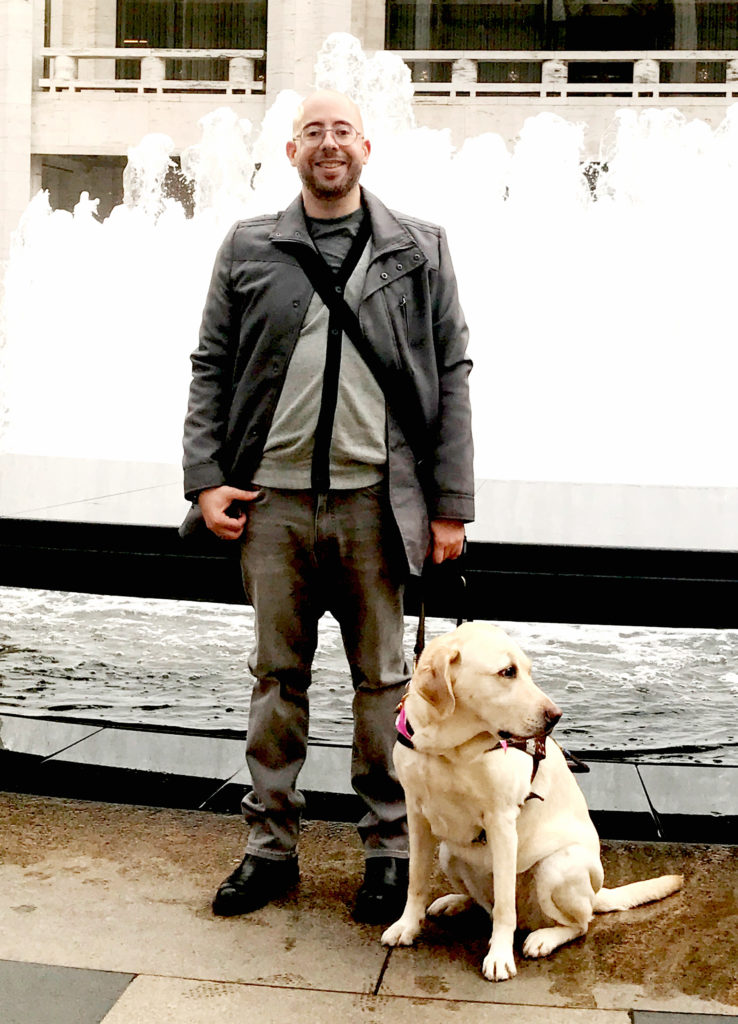 Gabriel smiling with his guide dog Posh in front of a large water fountain